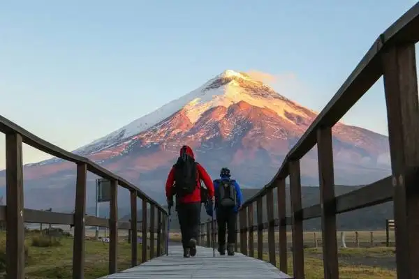 Excursionistas cruzan un puente de madera con el volcán Cotopaxi al amanecer