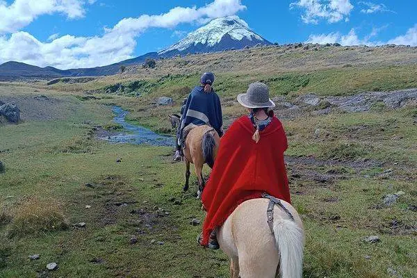 Two riders in ponchos cross a stream with snow-capped Cotopaxi rising in the distance
