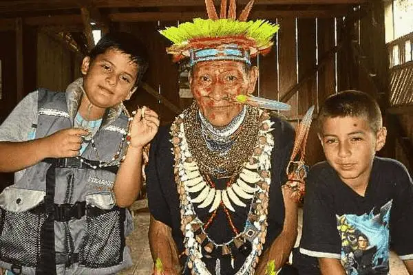 Indigenous elder in traditional feather headdress with two boys in a Cuyabeno community house