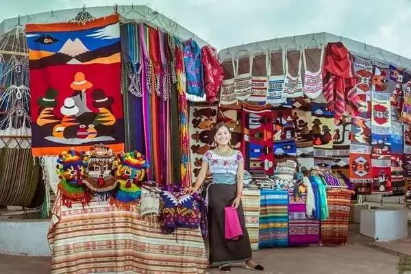 Otavalo Market vendor smiling beside stalls of colorful Andean textiles and blankets