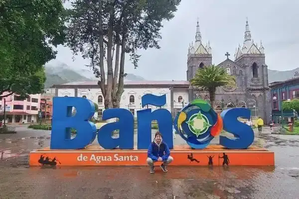 Tourist posing by the Banos de Agua Santa sign with the town church behind