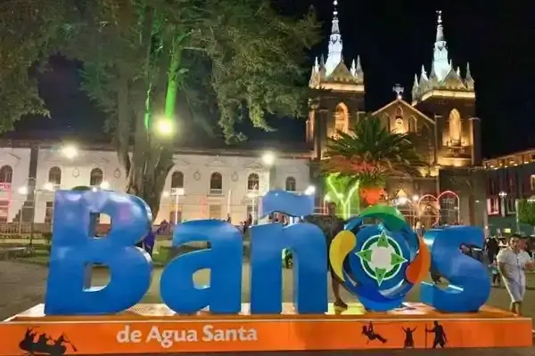 Illuminated Banos sign with the lit-up cathedral behind it in the town's main square at night