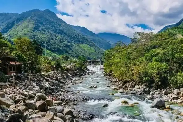 Rushing river through Banos town with green mountains and a small waterfall behind