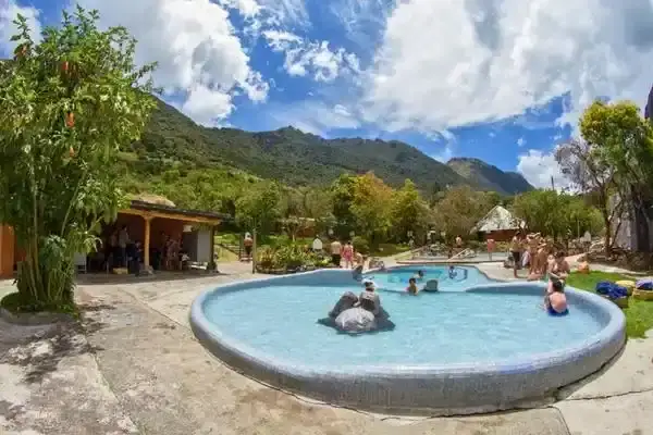 Visitors soaking in a large round thermal pool surrounded by green Andean mountains