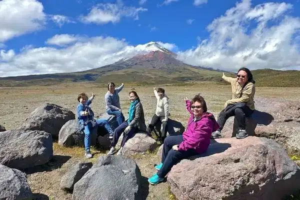Group of hikers posing on volcanic rocks with snow-capped Cotopaxi volcano behind them