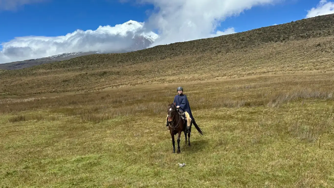 A rider in a poncho crosses the Cotopaxi paramo grassland with the volcano peeking through clouds